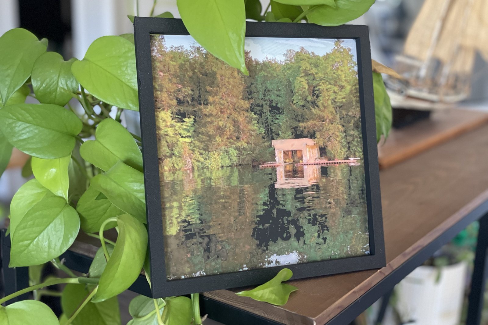 My Polychrome print of a cottage boathouse on a lake, framed on a shelf among the leaves of a houseplant