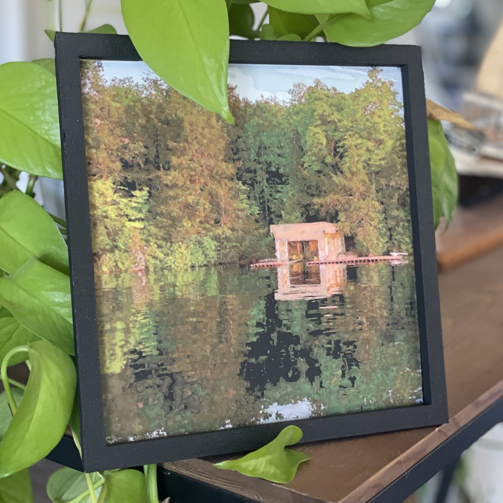 My Polychrome print of a cottage boathouse on a lake, framed on a shelf among the leaves of a houseplant